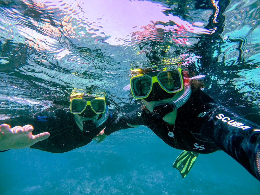 A beginner diver kneeling in shallow sand while an instructor holds the BCD strap during a supervised intro dive in Okinawa